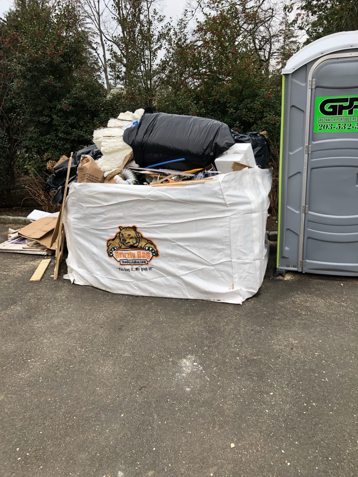 A Grizzly Bag flexible dumpster filled with household debris on a driveway, sized next to a portable restroom for scale.